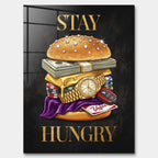 Nowoczesny obraz na szkle akrylowym przedstawiający burgera z banknotów, złota i zegarka oraz złoty napis STAY HUNGRY na czarnym tle. Wydruk galeryjny Huephant.
