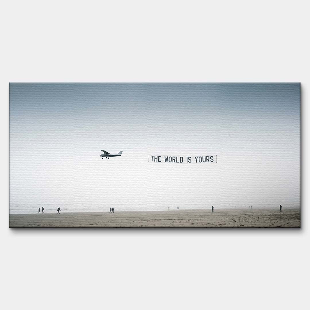 Minimalistyczny poziomy obraz na płótnie z awionetką ciągnącą napis The World Is Yours nad zamgloną plażą z sylwetkami ludzi, wydruk galeryjny.