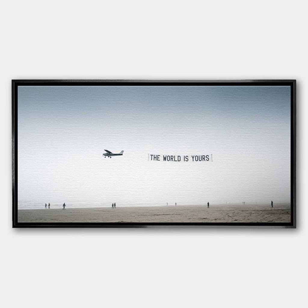 Minimalistyczny poziomy obraz na płótnie z awionetką ciągnącą napis The World Is Yours nad zamgloną plażą z sylwetkami ludzi, wydruk galeryjny.