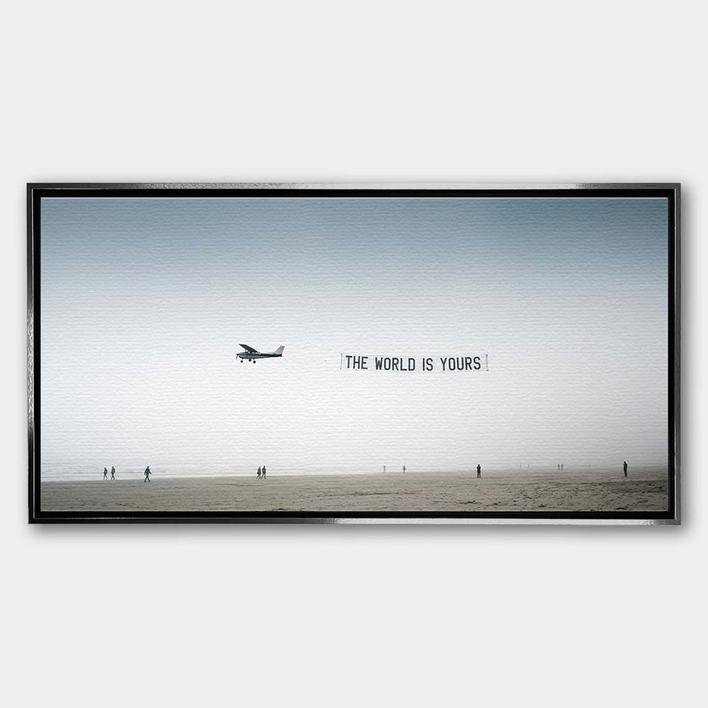 Minimalistyczny poziomy obraz na płótnie z awionetką ciągnącą napis The World Is Yours nad zamgloną plażą z sylwetkami ludzi, wydruk galeryjny.