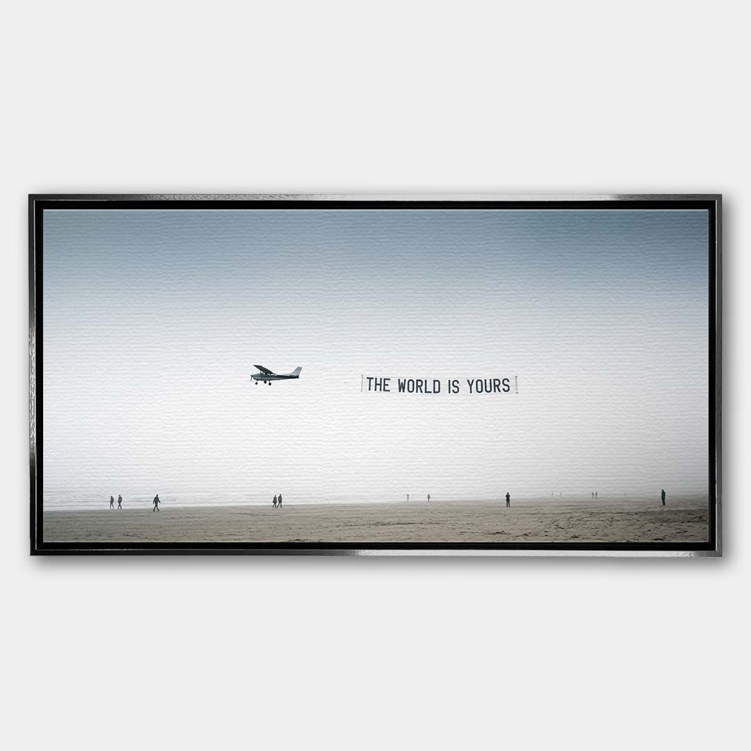 Minimalistyczny poziomy obraz na płótnie z awionetką ciągnącą napis The World Is Yours nad zamgloną plażą z sylwetkami ludzi, wydruk galeryjny.