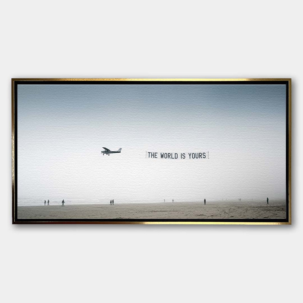 Minimalistyczny poziomy obraz na płótnie z awionetką ciągnącą napis The World Is Yours nad zamgloną plażą z sylwetkami ludzi, wydruk galeryjny.